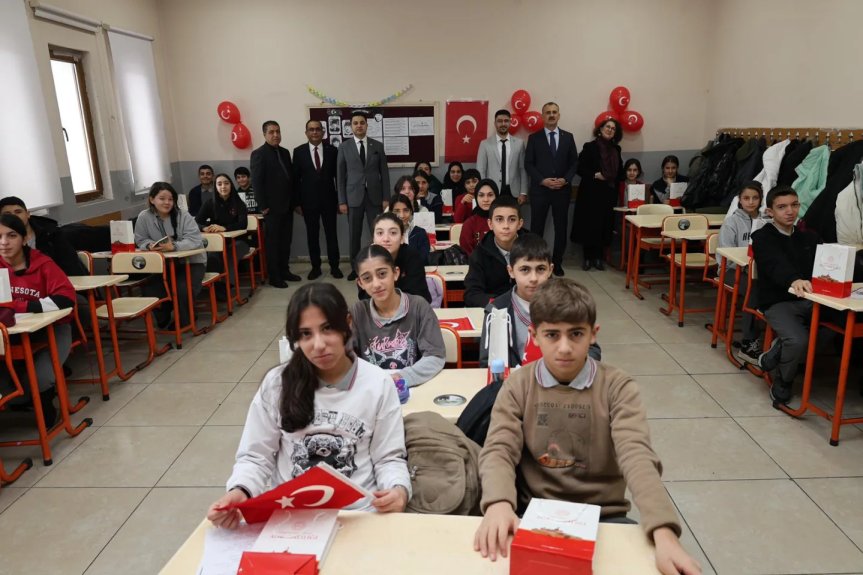 Sınıfta sıralarında oturan öğrenciler ve arka planda okul yöneticilerinin yer aldığı kalabalık sınıf ortamı